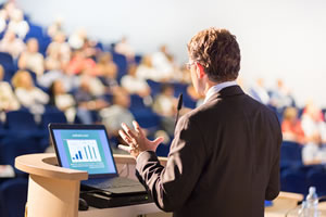Photo of a male lecturer in a business suit standing behind a lecturn on a stage speaking to a large audience