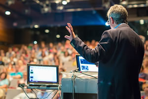 Photo of a male lecturer in a business suit speaking to a large audience in an auditorium